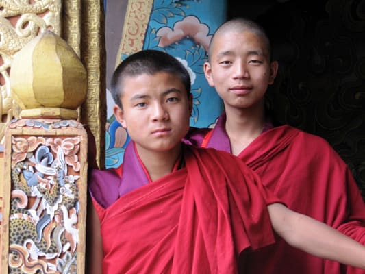 Two young monks in red robes standing next to a wall.