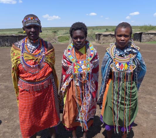 Three women in colorful dresses standing on a dirt field.