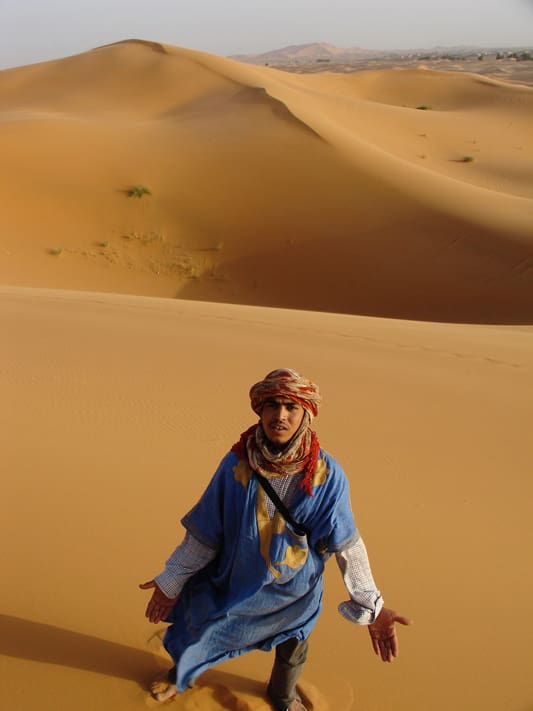 A man in blue and red standing on top of a sand dune.
